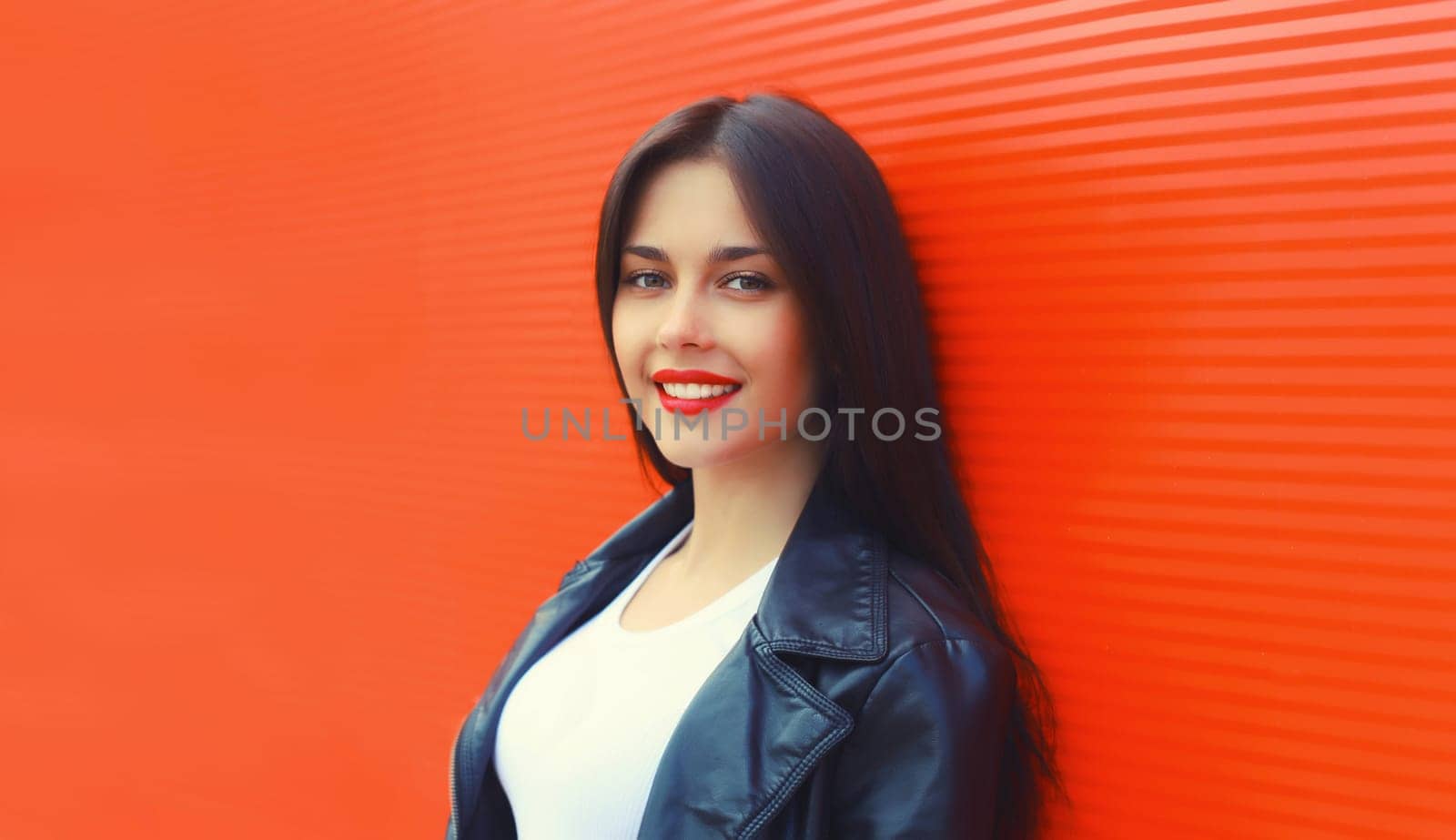 Portrait of beautiful brunette smiling young woman model posing in black leather jacket on red background