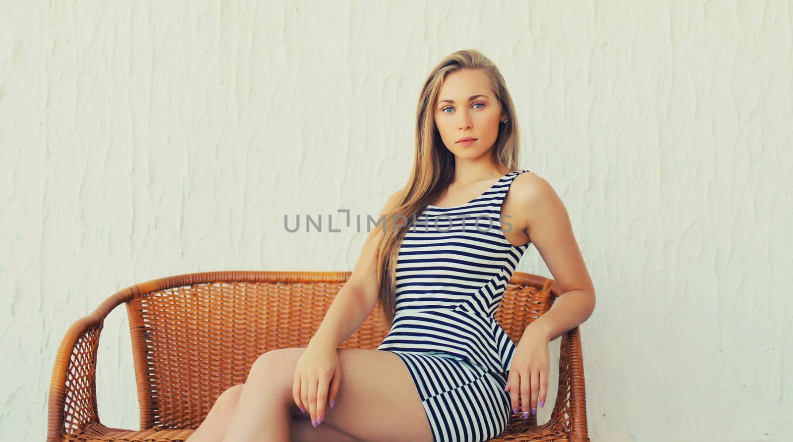 Portrait of beautiful blonde young woman in summer striped dress resting on armchair