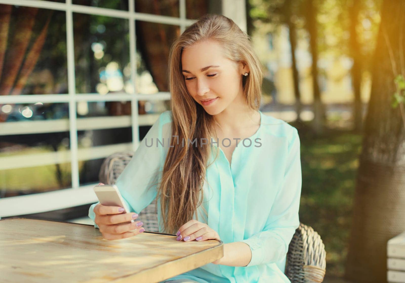 Happy caucasian young woman 20s with mobile phone looking at device while sitting at the table in street cafe in summer park