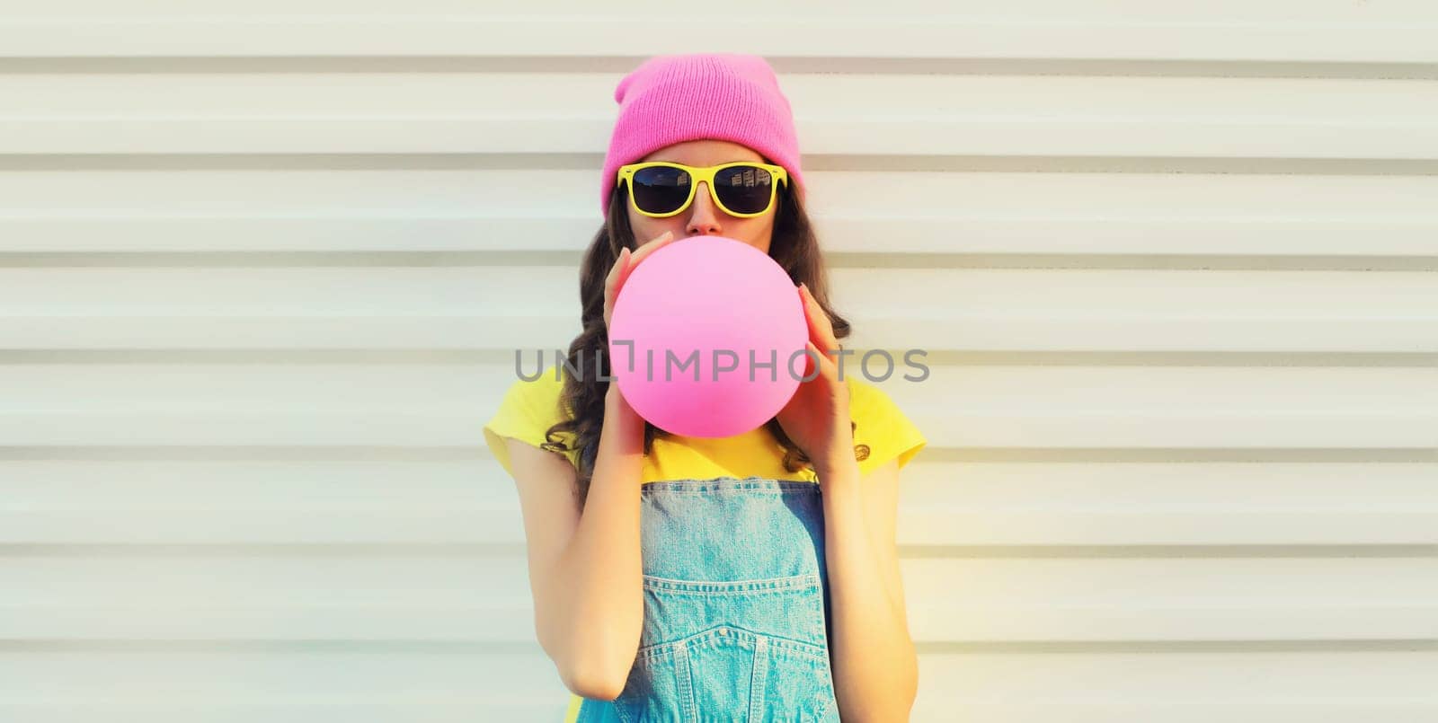 Fashion portrait of stylish cool young woman inflating chewing gum or balloon in colorful clothes, pink hat, yellow sunglasses on white background