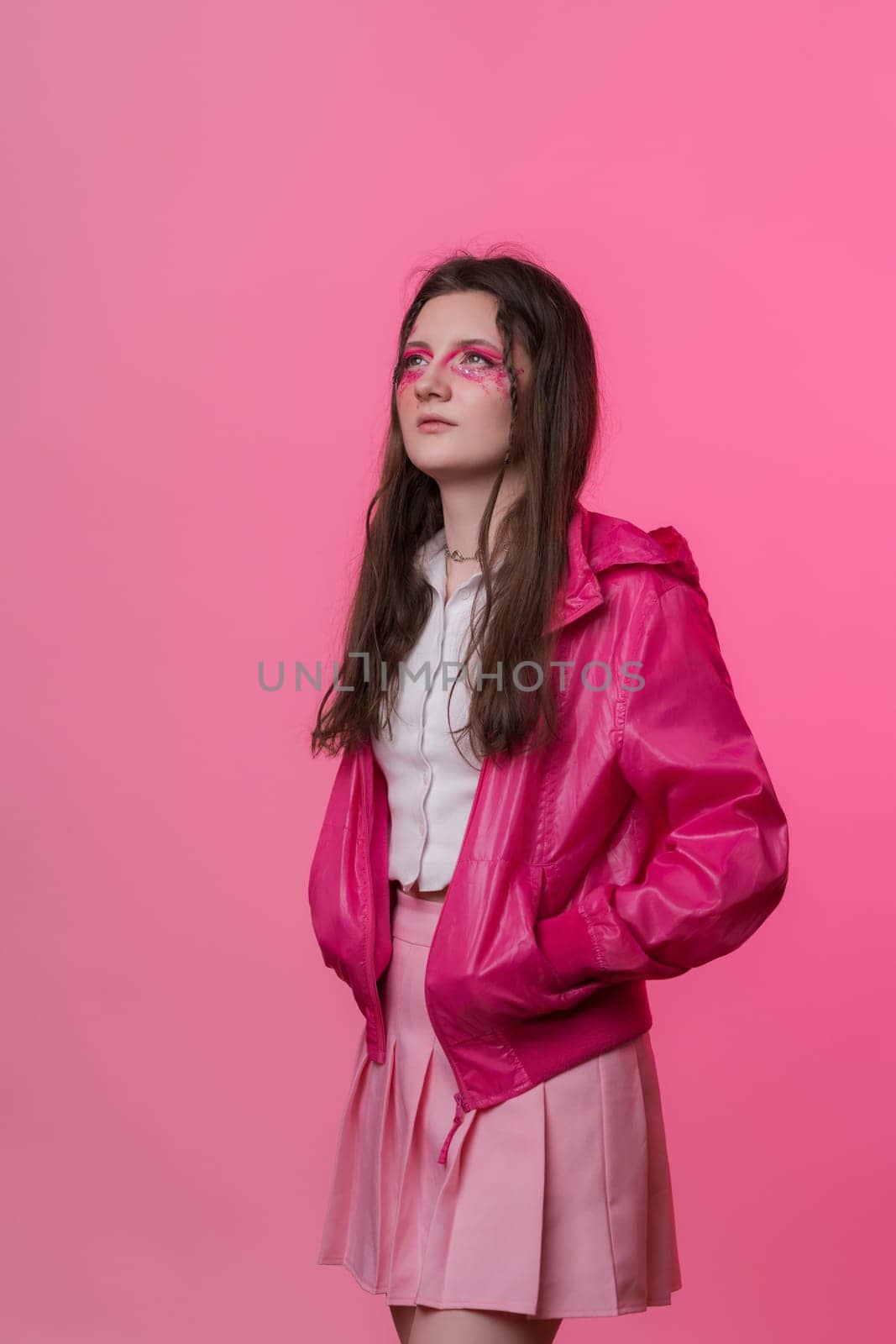 Portrait of woman with pink make-up with sequins and arrows, dressed in pink jacket, skirt and white t-shirt. Young adult Caucasian ethnicity hipster female on pink background. Part of photo series