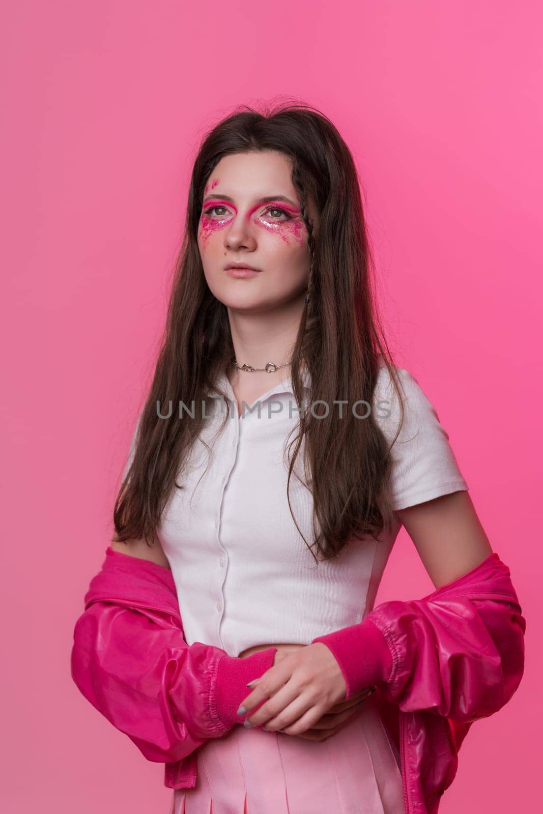 Portrait of female with pink make-up with sparkles and arrows, dressed in pink jacket, skirt and white t-shirt. Studio shot of pretty hipster Caucasian woman on pink background. Part of photo series