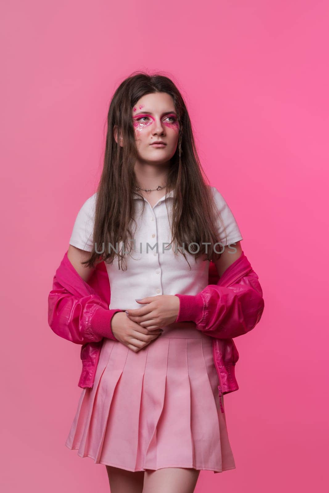 Portrait of serious woman with pink make-up with sparkles and arrows, dressed in pink jacket, skirt, white t-shirt. Studio shot of young adult hipster female on pink background. Part of photo series