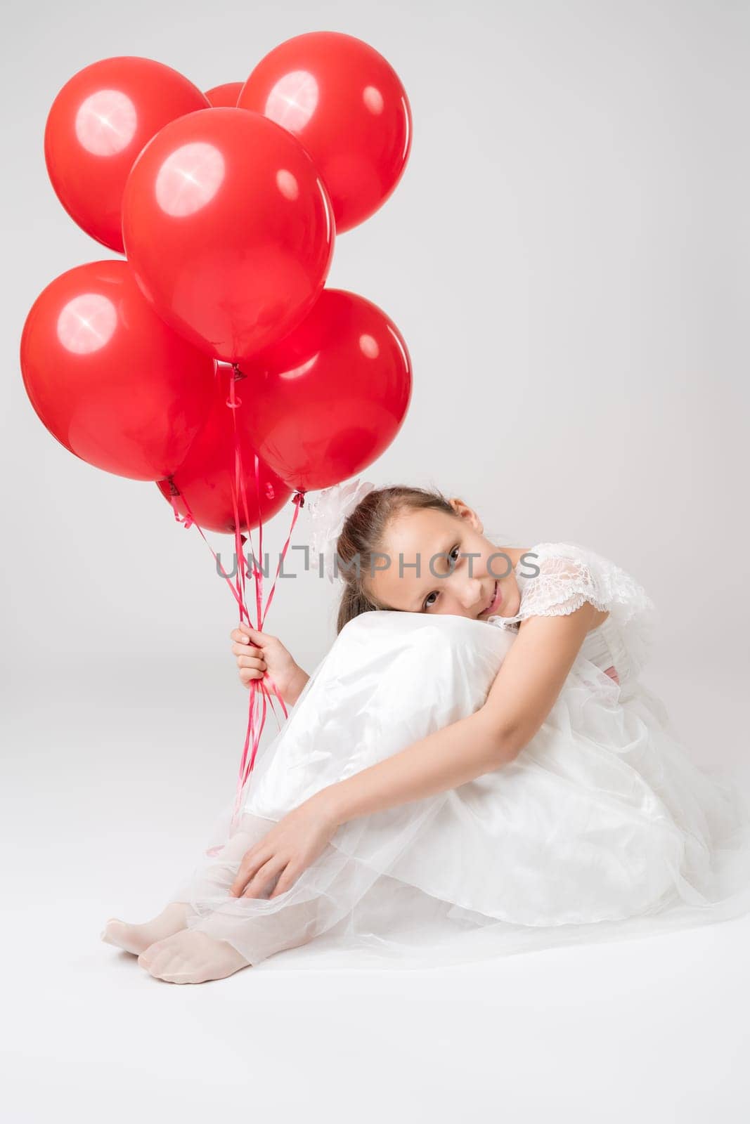 Romantic girl in white dress holding lot of red balloons in hand, put head on her knees, sitting on white background and smiles looking at camera. Studio shot. Part of photo series.