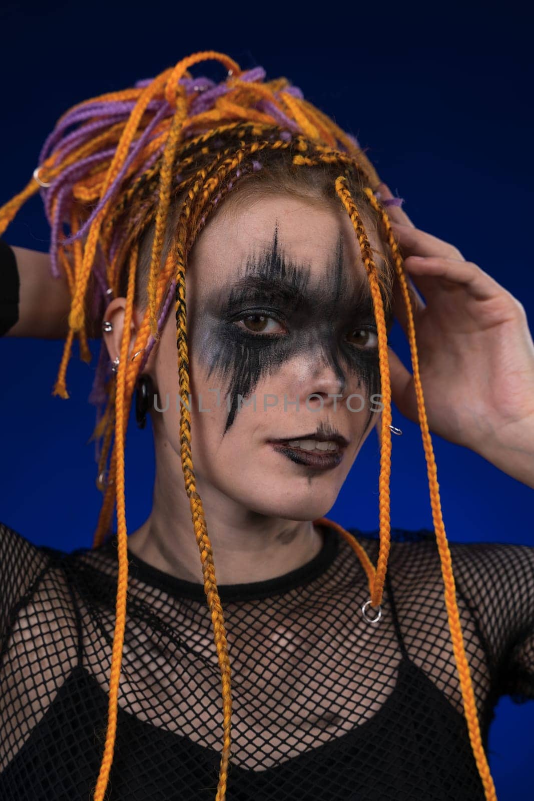 Informal young woman with colored dreadlocks hairstyle and horror black stage make up painted on face. Studio shot on blue background. Part of photo series