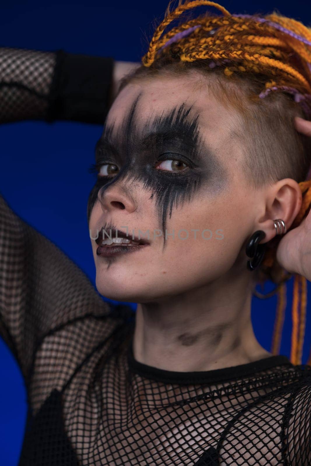 Headshot of young woman with horror black stage make up painted on face and colored without dangerous dreadlocks hairstyle. Studio shot cinematic portrait on blue background. Part of photo series