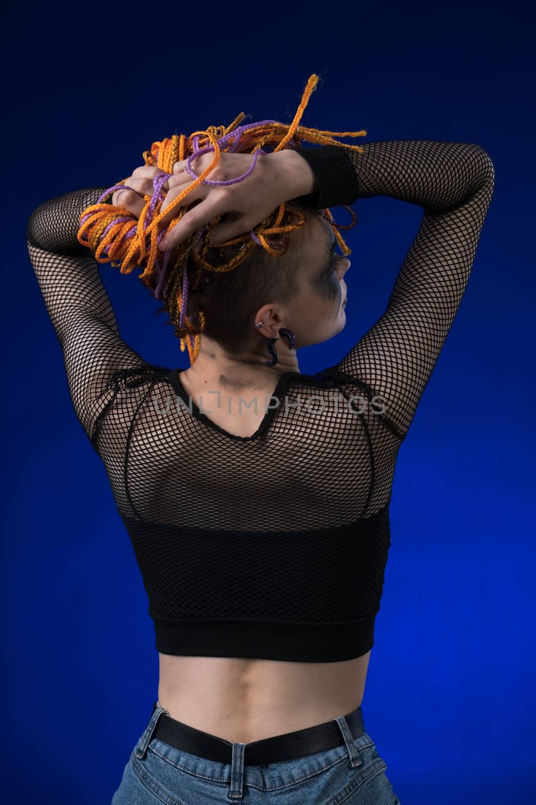 Rear view of young woman with colored braids hairdo and horror black stage make-up painted on face. Studio shot on blue background. Part of photo series