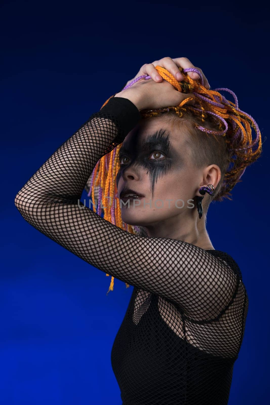 Shy young female with orange color braids hairdo and horror black stage makeup painted on face. Studio shot on blue background. Part of photo series