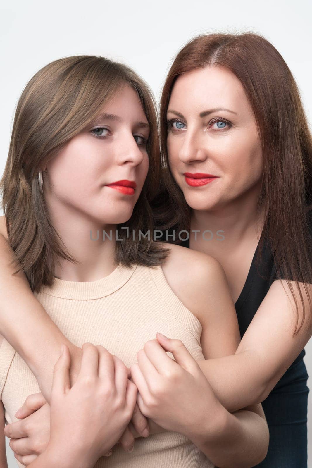 Adult mother gently hugging her teenage daughter standing behind. Friendly Caucasian ethnicity daughter and mom with gray eyes and long hair. Togetherness concept. Studio shot, part of photo series