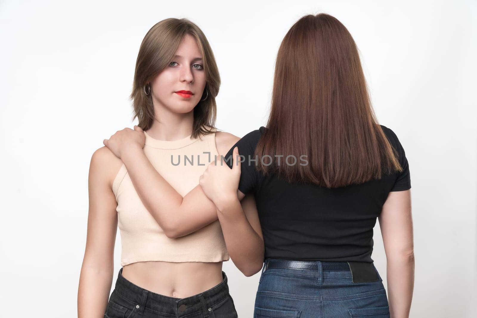 Two generations, teenager daughter and adult mother hugging each other. Caucasian ethnicity daughter stands facing and looking at camera, mom standing with her back. Studio shot, part of photo series