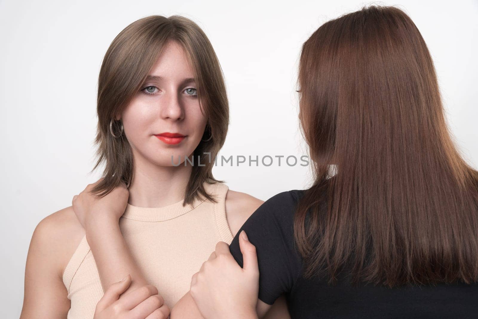 Portrait of daughter with gray eyes and long hair standing facing, looking at camera, mother stands with her back. Two generations, teenager daughter and adult mom hugging each other. Part of series