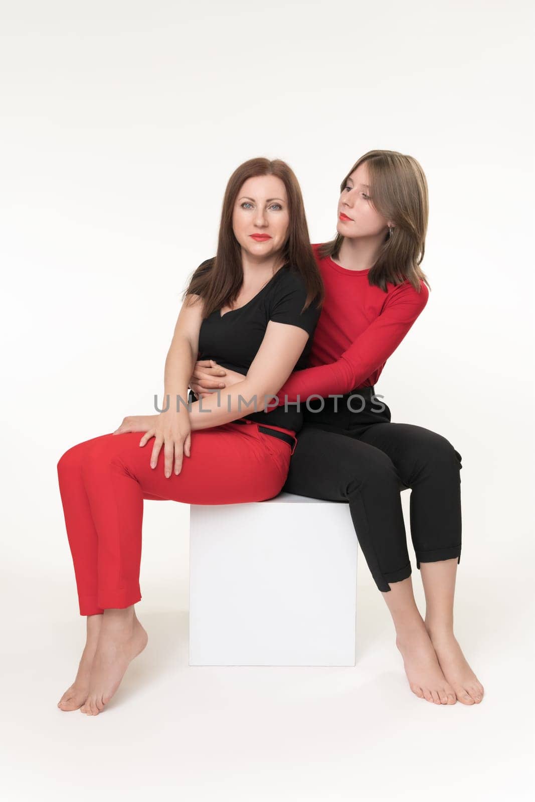 Mom and daughter sitting on white cube box on white background. Teenager daughter hugging mother and looking at her. Happy mommy looking at camera. Togetherness concept. Part of photo series