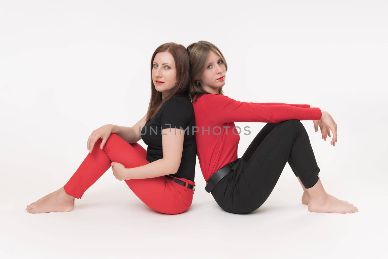 Mother and teenager daughter sitting with their backs to each other and sideways to camera on white background. Both females seriously looking at camera. Studio shot, part of photo series