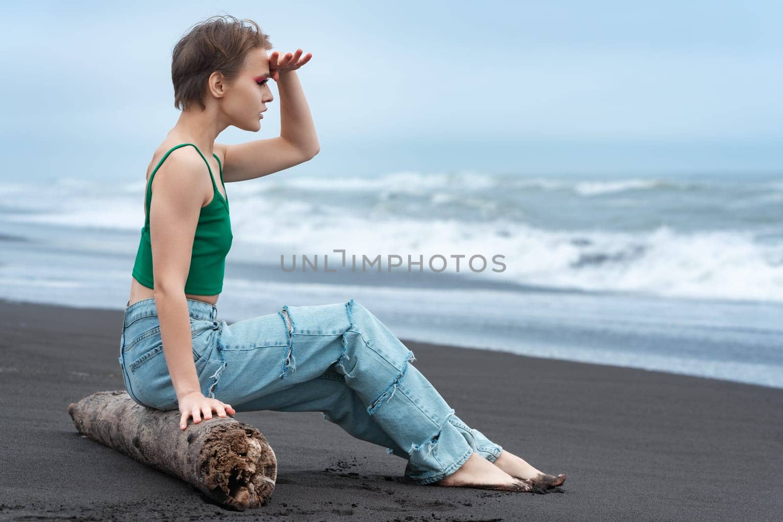 Hipster woman sitting on log on black sandy beach of Pacific Ocean, raised her hand to forehead and looking at view at breaking sea waves. Blonde woman with short hair wearing green top and blue jeans
