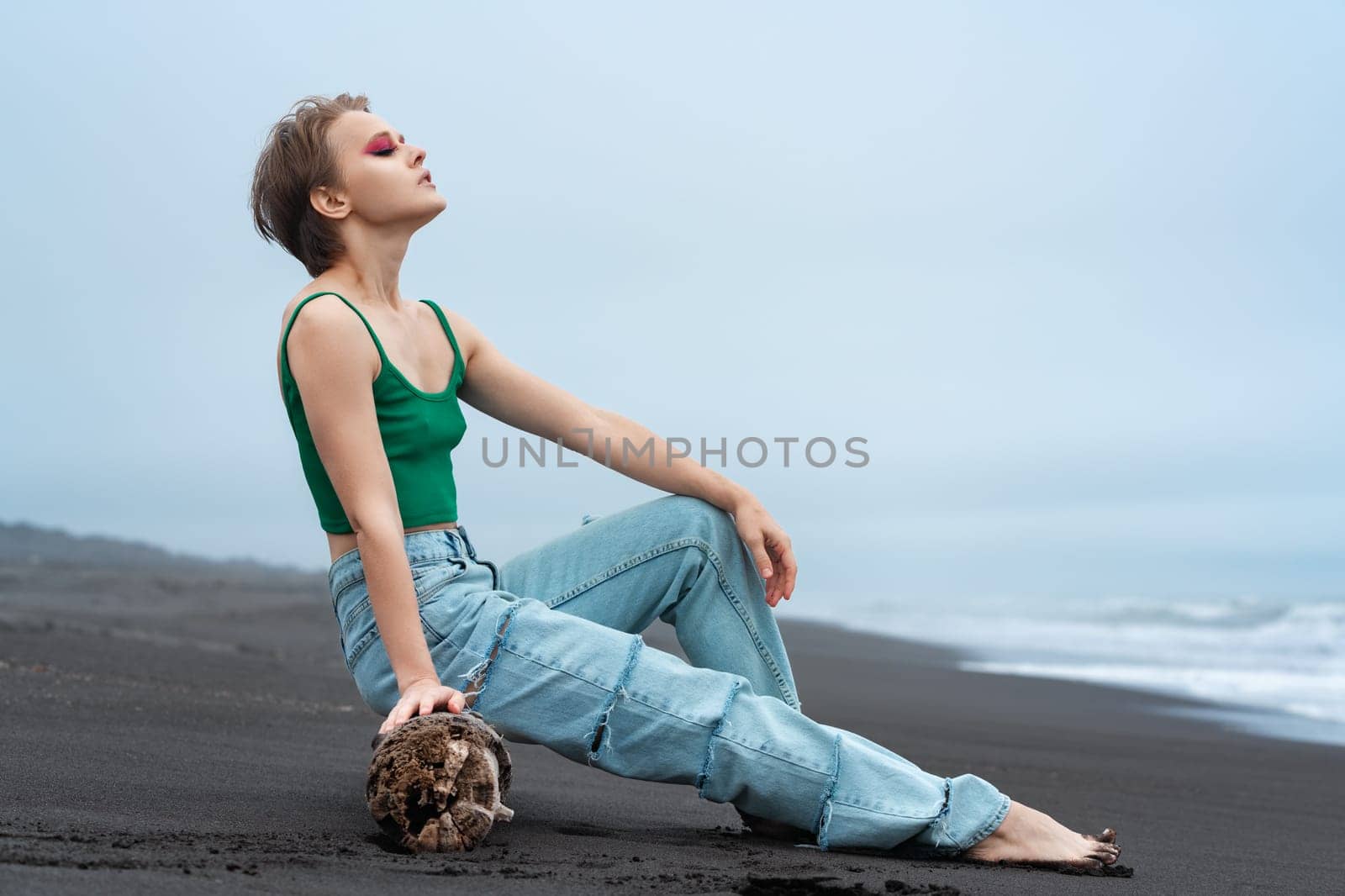 Awesome hipster woman with eyes closed and relaxed expression looks like ultimate millennial tourist. Blonde wearing green top and blue jeans, leaning back on log on black sandy beach of Pacific Ocean