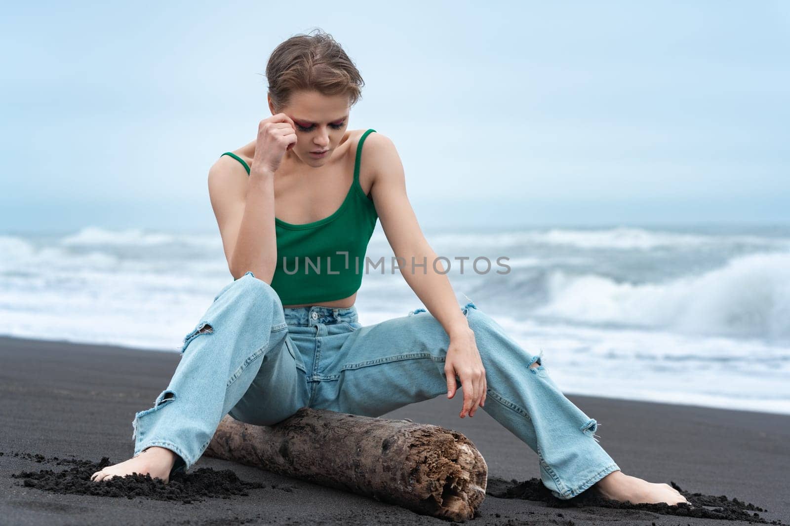 Blonde woman sitting on log with her legs spread and head lowered thoughtfully, looking down on black sandy beach of Pacific Ocean. Millennial woman with short hair wearing blue jeans and green top