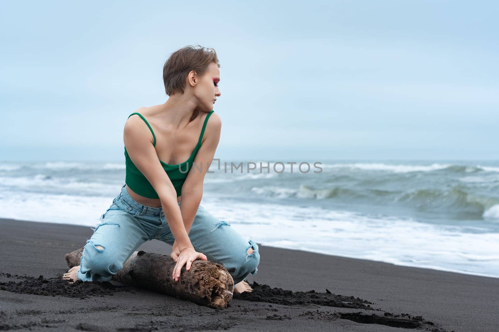 Sexy blonde woman striking playful pose on log, sitting like she riding horse and turning head to side with flirty eyes closed. Hipster woman relaxing on beach of Pacific Ocean during summer vacation