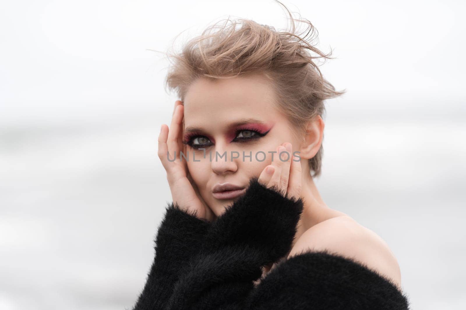Blonde woman posing outside against white natural background and her bright makeup really makes her stand out. Sensuality woman looking at camera with head in hands and looks absolutely stunning