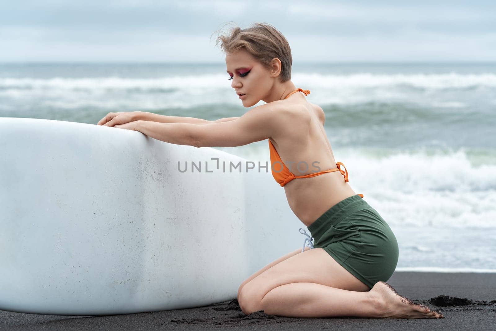 Female surfer, with her eyes closed, is kneeling and holding her surfboard during summer vacation. Sportswoman is wearing bikini top and shorts, and appears to exude sensuality