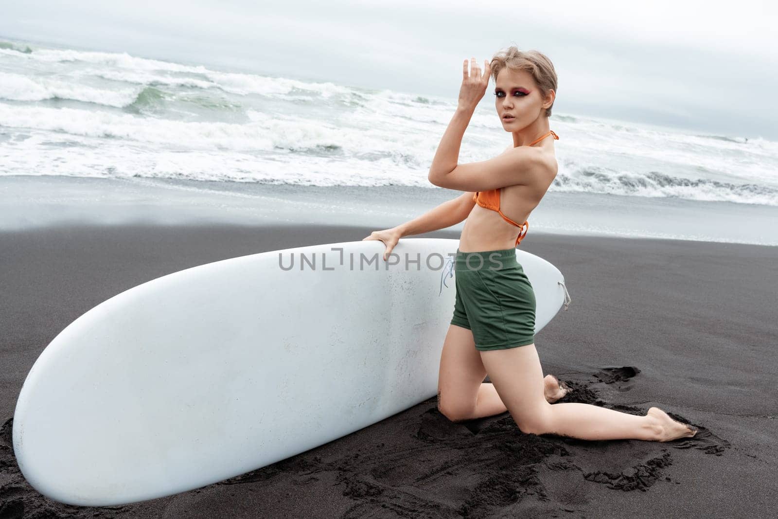 Female surfer kneeling on black sand of beach, holding her surfboard in hands and looking at camera. Sportswoman clearly skilled and passionate rider who has great deal of experience and knowledge
