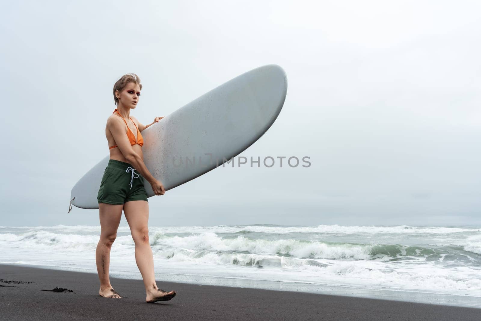 Woman surfer walks on black sand beach carrying white surfboard against backdrop of sea waves during summer vacation. Sporting woman in bikini top, shorts and barefoot. Physical activity concept