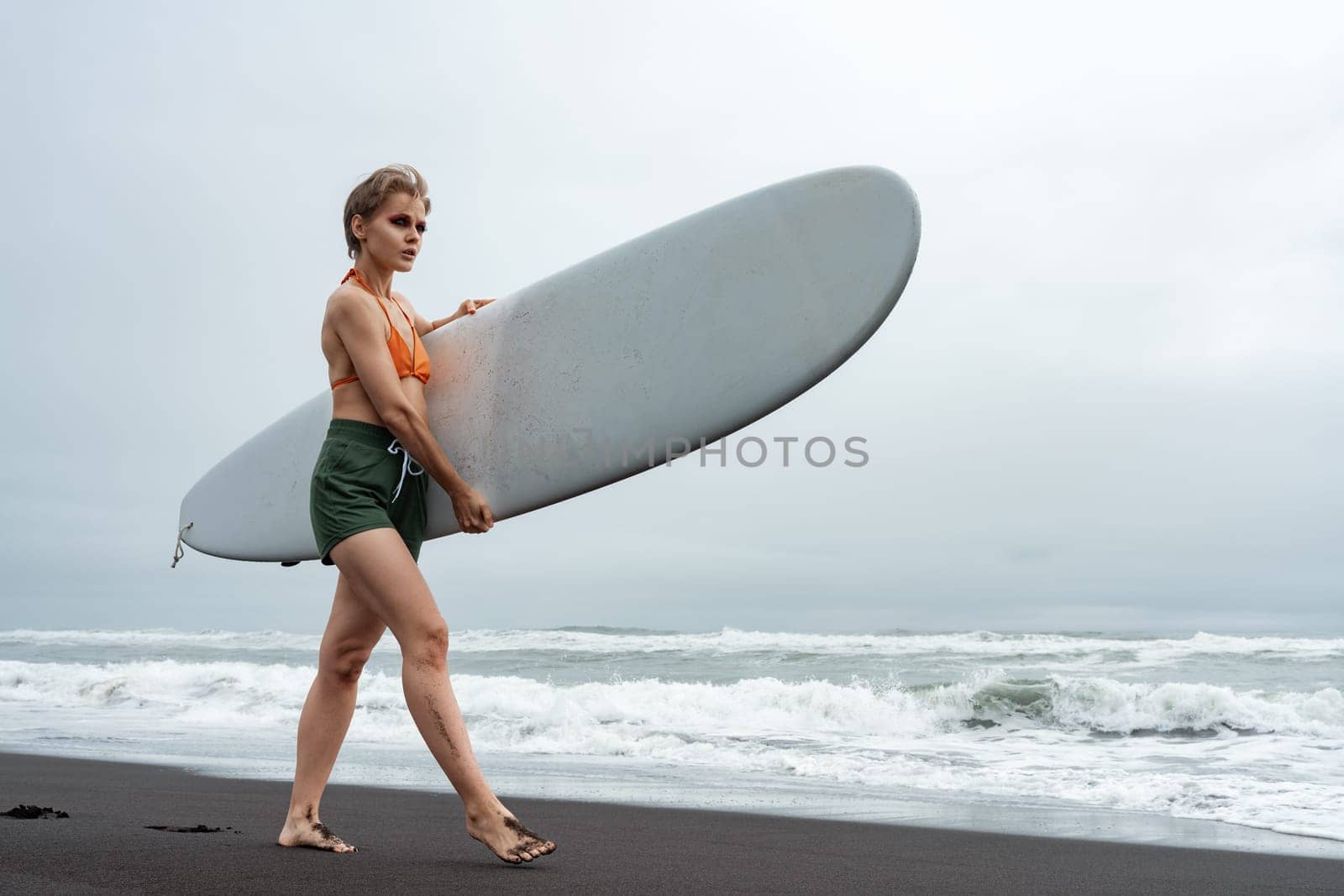 Sporty surfer is walking barefoot on black sand beach during summer holiday, carrying white surfboard against background of sea waves. Female sportswoman wearing bikini top, shorts and barefoot