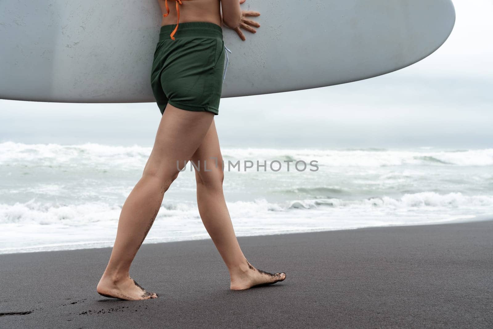 Surfer woman carrying surfboard walking along beach with sea waves in background. Side view of sensuality female legs and buttocks is an athletic figure. Concept of extreme sports