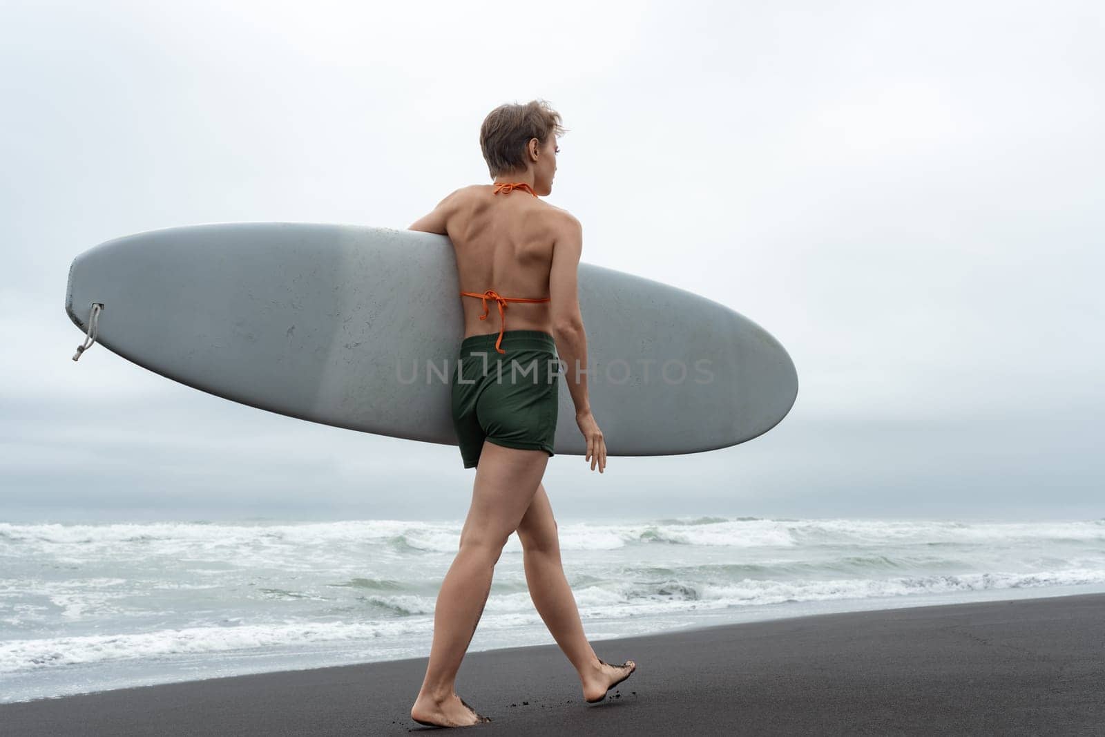 Rear view of sexuality female surfer walking on black sandy beach carrying white surfboard against background of ocean waves during summer resort holiday. Healthy lifestyle concept