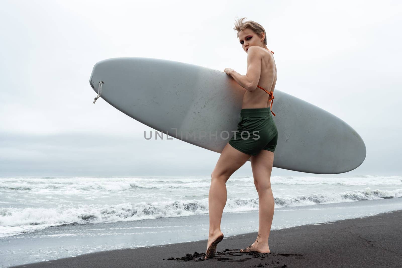 Female surfer standing on black sandy beach holding white surfboard against background of sea waves during summer beach vacation. Sporty woman looking over her shoulder. Active lifestyle concept