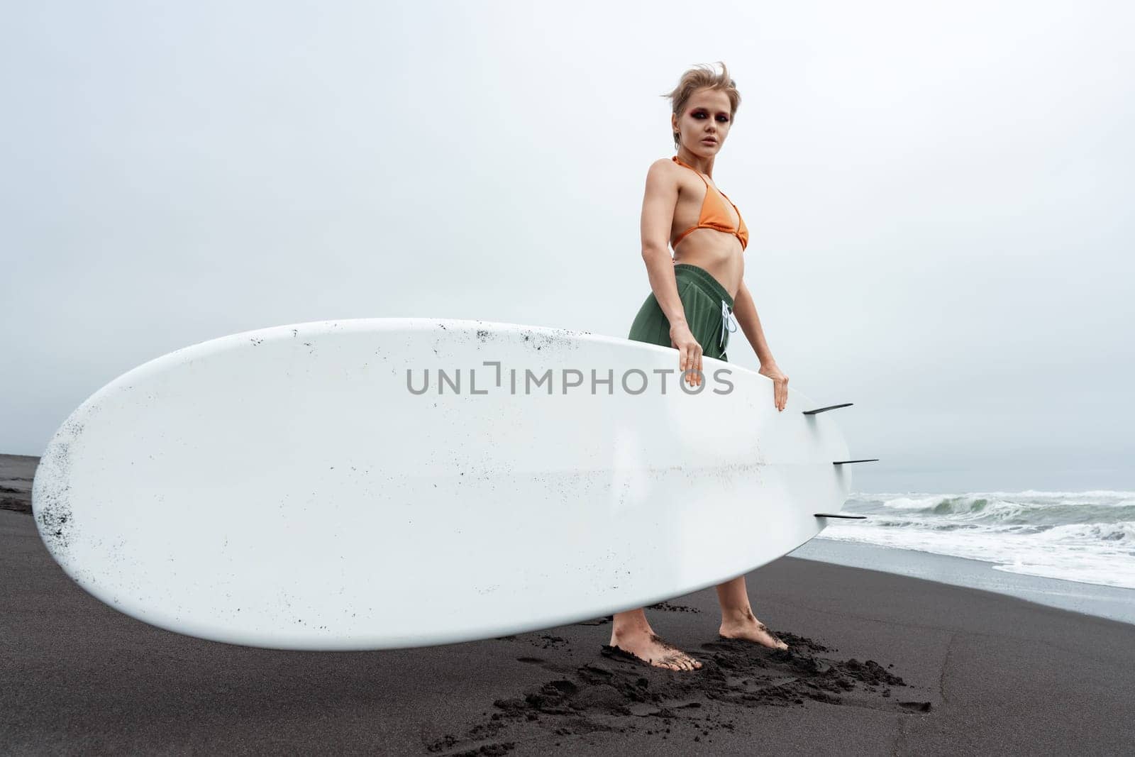 Female surfer standing on black sandy beach holding surfboard against background of sea waves during summer beach holiday. Woman looking at camera. Active lifestyle concept. Full length, wide shot