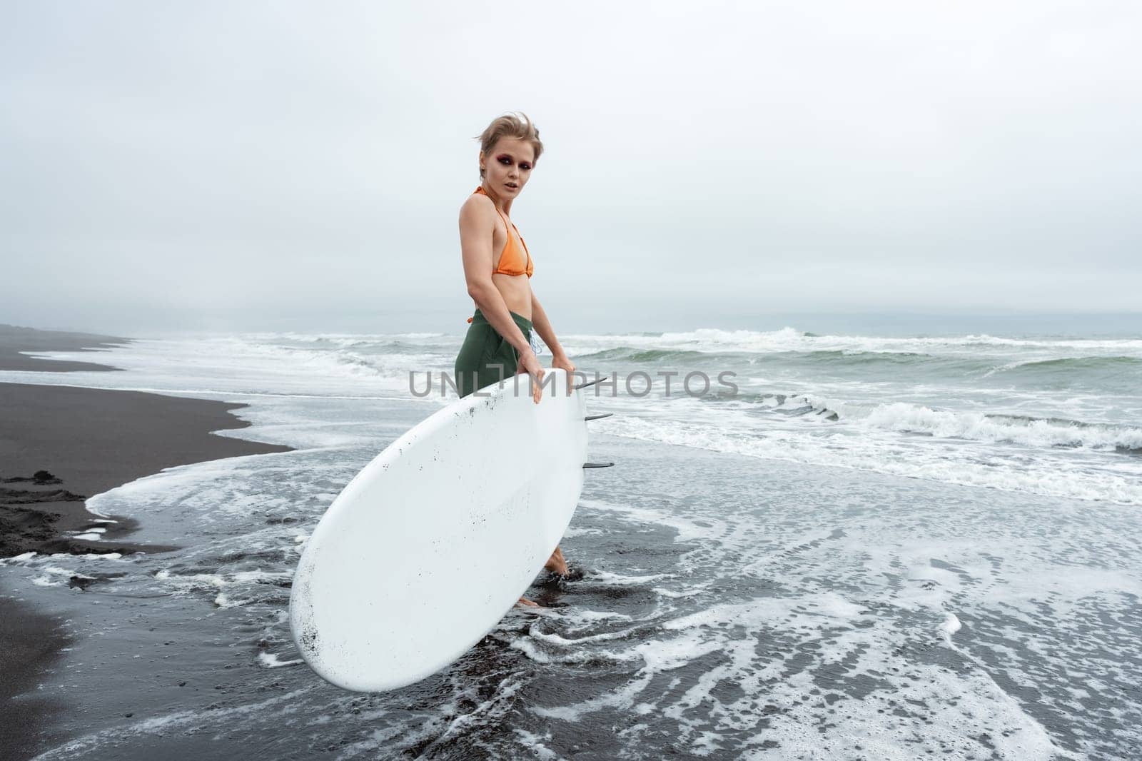 Woman surfer walking in ankle deep water of sea waves on beach carrying white surfboard against background of ocean during summer beach holiday. Sportswoman looking at camera. Full length, wide shot