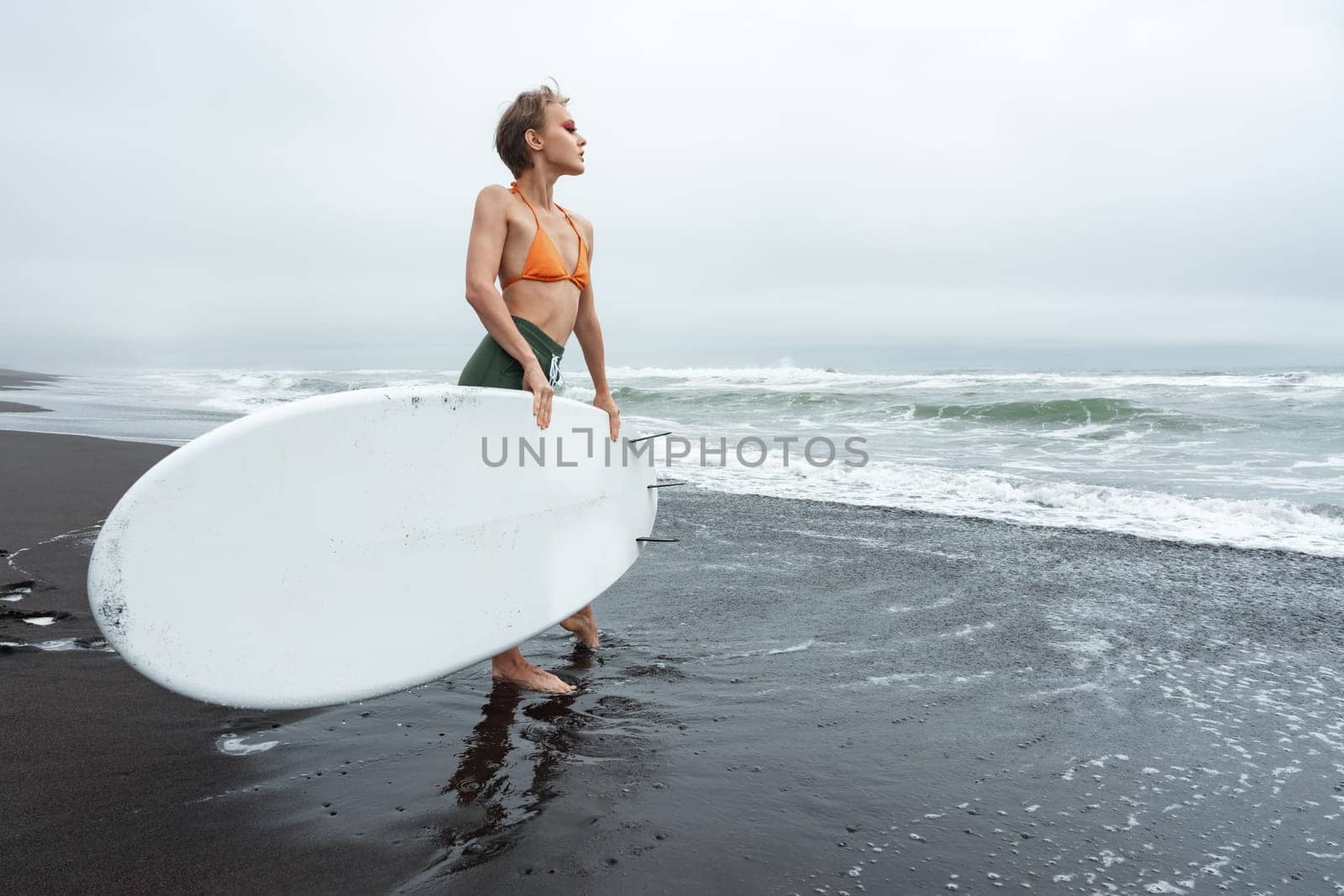 Attractive female surfer standing on black sandy beach holding white surfboard during summer beach holiday. This is perfect image to capture excitement and beauty of surfing. Full length, wide shot