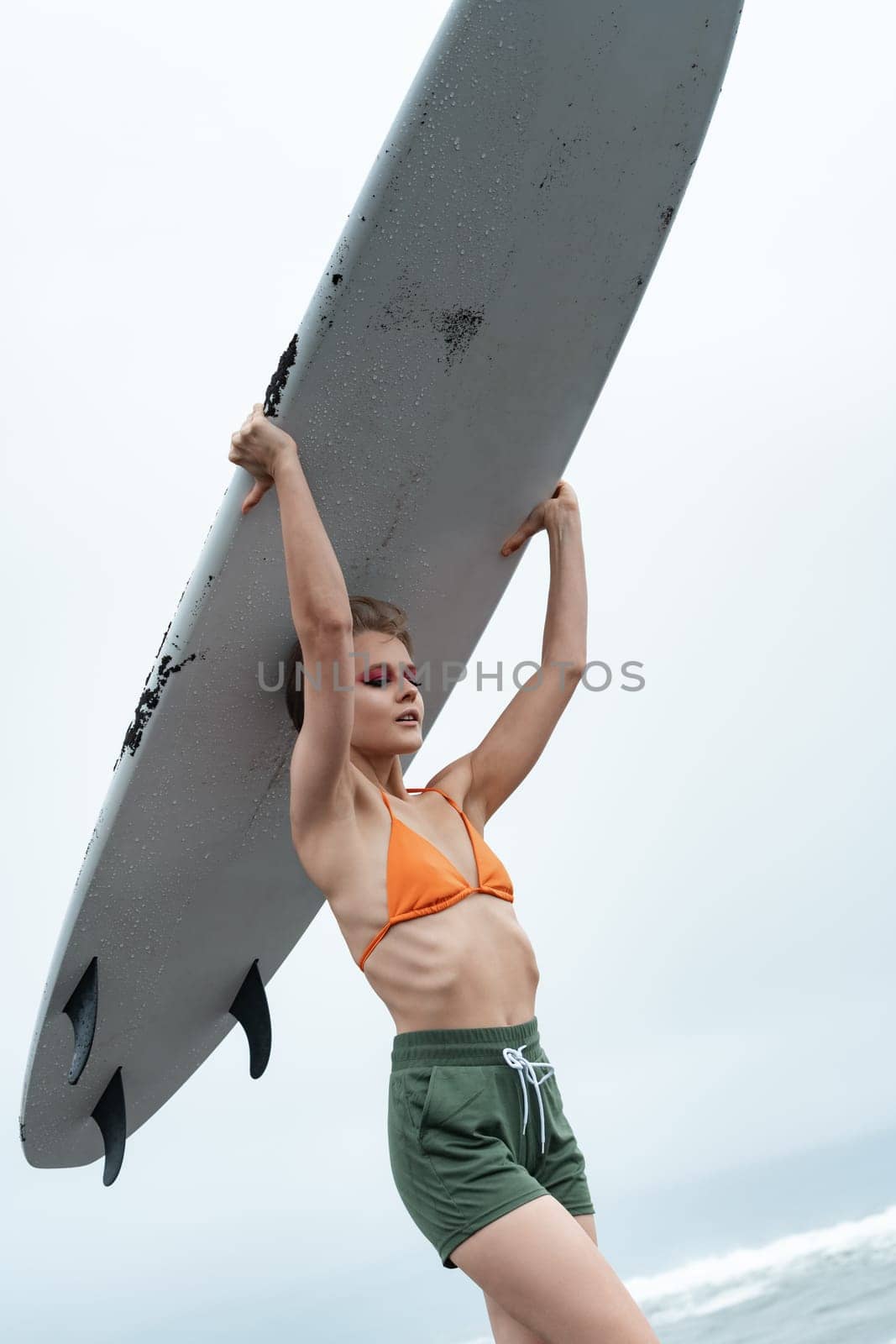 Sexuality female surfer carrying white surfboard on head during sports training. Beauty athletic woman in bikini top, shorts. Close-up Dutch angle view, low angle shot. Concepts of sport activities