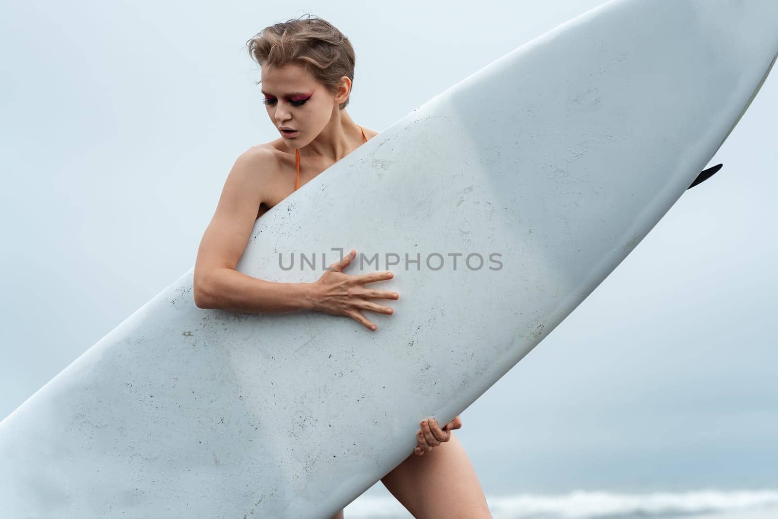 Close-up view of female sports fashion model holding white surfboard and standing behind it. Woman surfer in relaxed pose, looking down during summer vacation. Concept of sports and active lifestyle