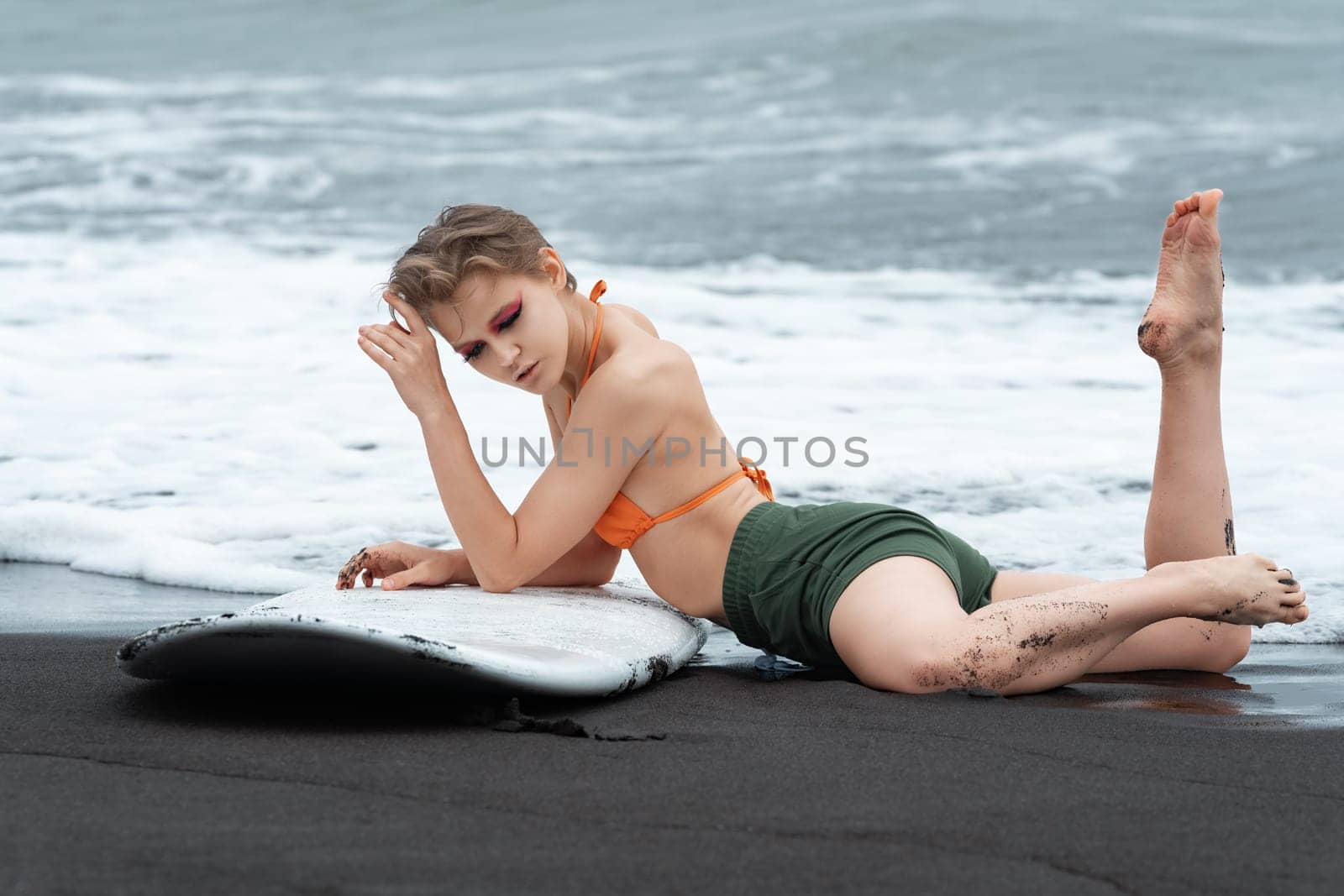 Dreamy sexy surfer is lying on black sand of Pacific Ocean beach with surfboard, showing off fit and athletic body. Sexuality sportswoman relaxing after sports training during summer beach vacation