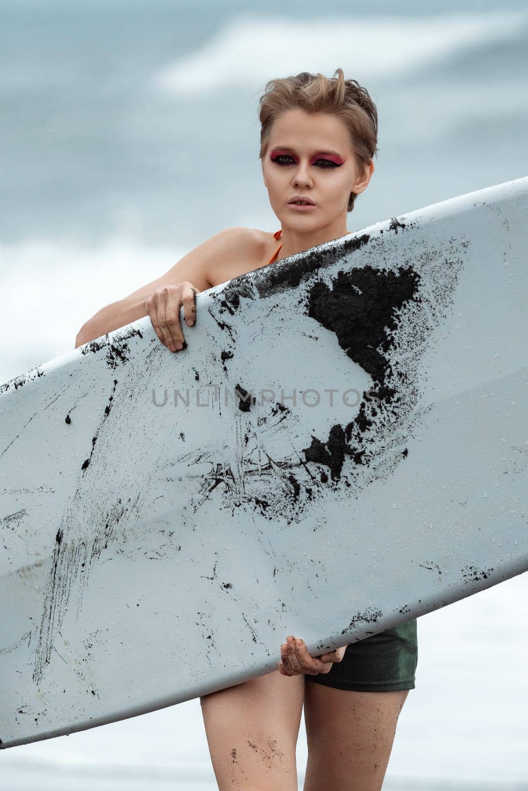 Female surfer carrying white surfboard is walking on beach and looking at camera with sea waves in background. Beauty woman doing sports symbolizes concept of physical activity and healthy lifestyle