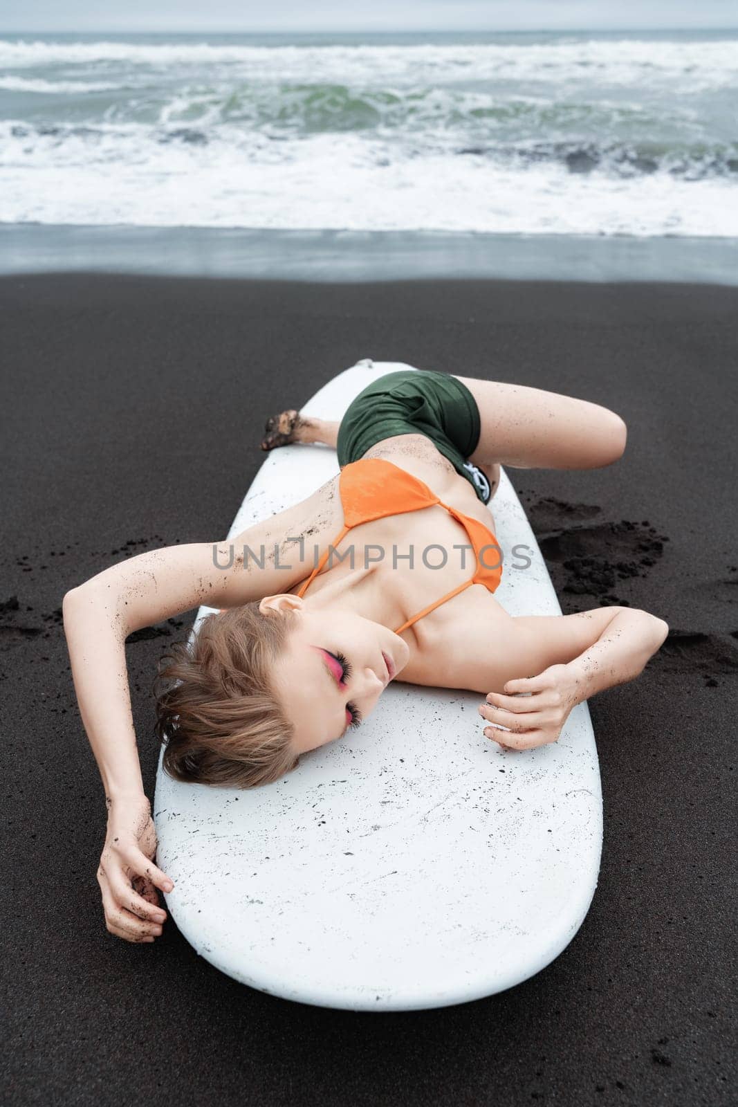 Female surfer is lying on white surfboard on black sandy beach in Pacific Ocean. With her eyes closed and in relaxed pose, she is taking well deserved break after an intense sports training session