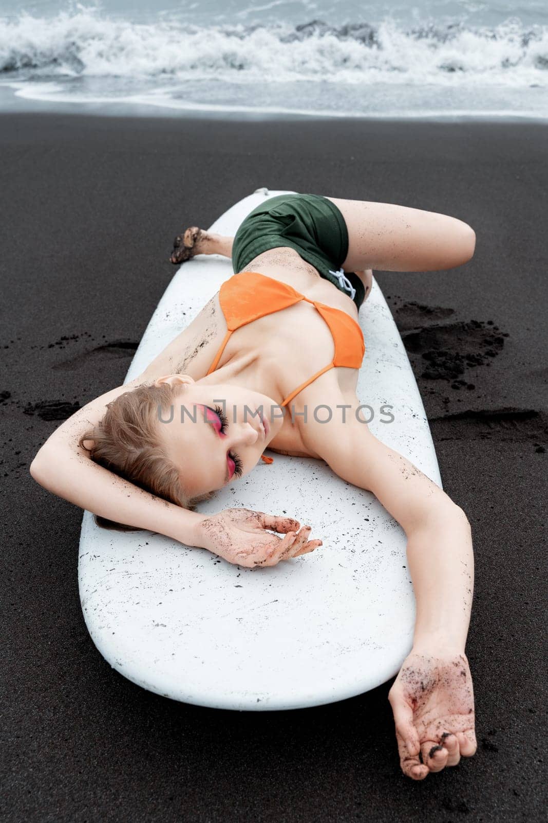 Female surfer is enjoying well deserved break on black sandy beach, lying on white surfboard with eyes closed. Sensuality sports fashion model looks so relaxed after an intense sports training session