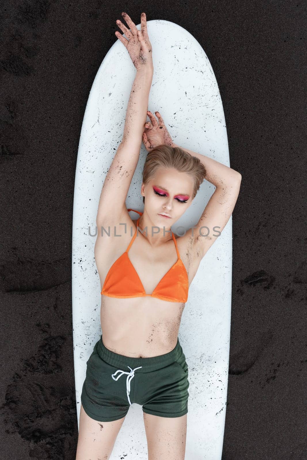 Top view of slim female surfer looks so relaxed and at peace, lying on surfboard on black sandy beach. Sensuality sports fashion model in bikini top and shorts, making her look both sexy and athletic