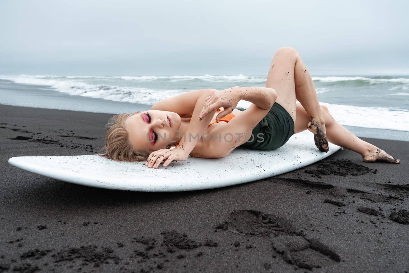 Female surfer is lying on white surfboard on black sandy beach. Sports fashion model with closed eyes is posing in bikini top and shorts, showing off her fit and athletic body after an intense workout
