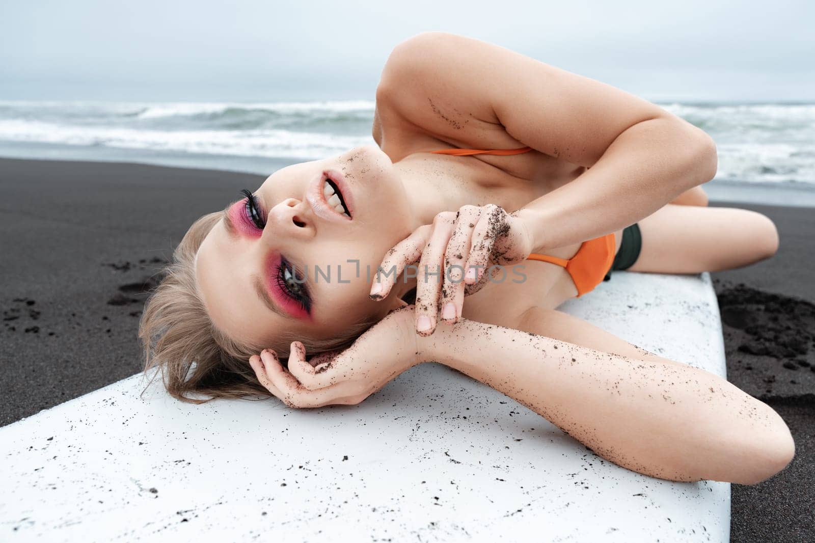 Woman surfer with bright makeup lying on surfboard on black sand beach on ocean shore. Close-up view really captures beauty of moment. Sensuality sports fashion model looks so relaxed and happy