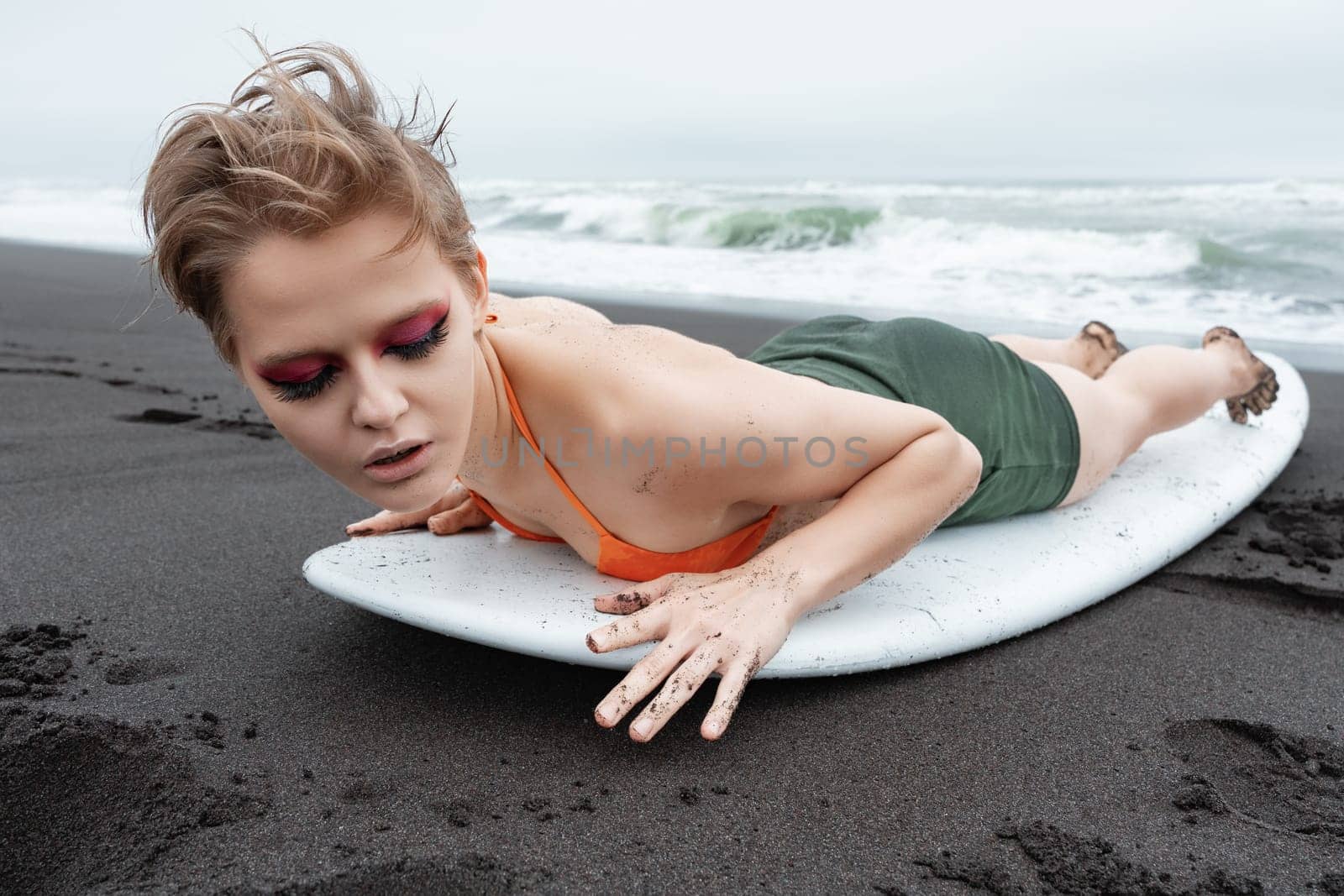 Blonde surfer woman with bright makeup looks stunning as she lies on front of surfboard on sandy beach. With her eyes closed, surfer is fully immersed in moment, feeling rush of sea and warmth of sun