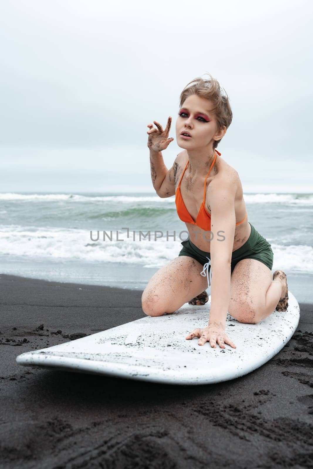 Sensuality woman surfer strikes pose on her knees atop white surfboard. Sports fashion model looking at camera. Sportswoman in bikini top and shorts, embodying combination of athleticism and beauty