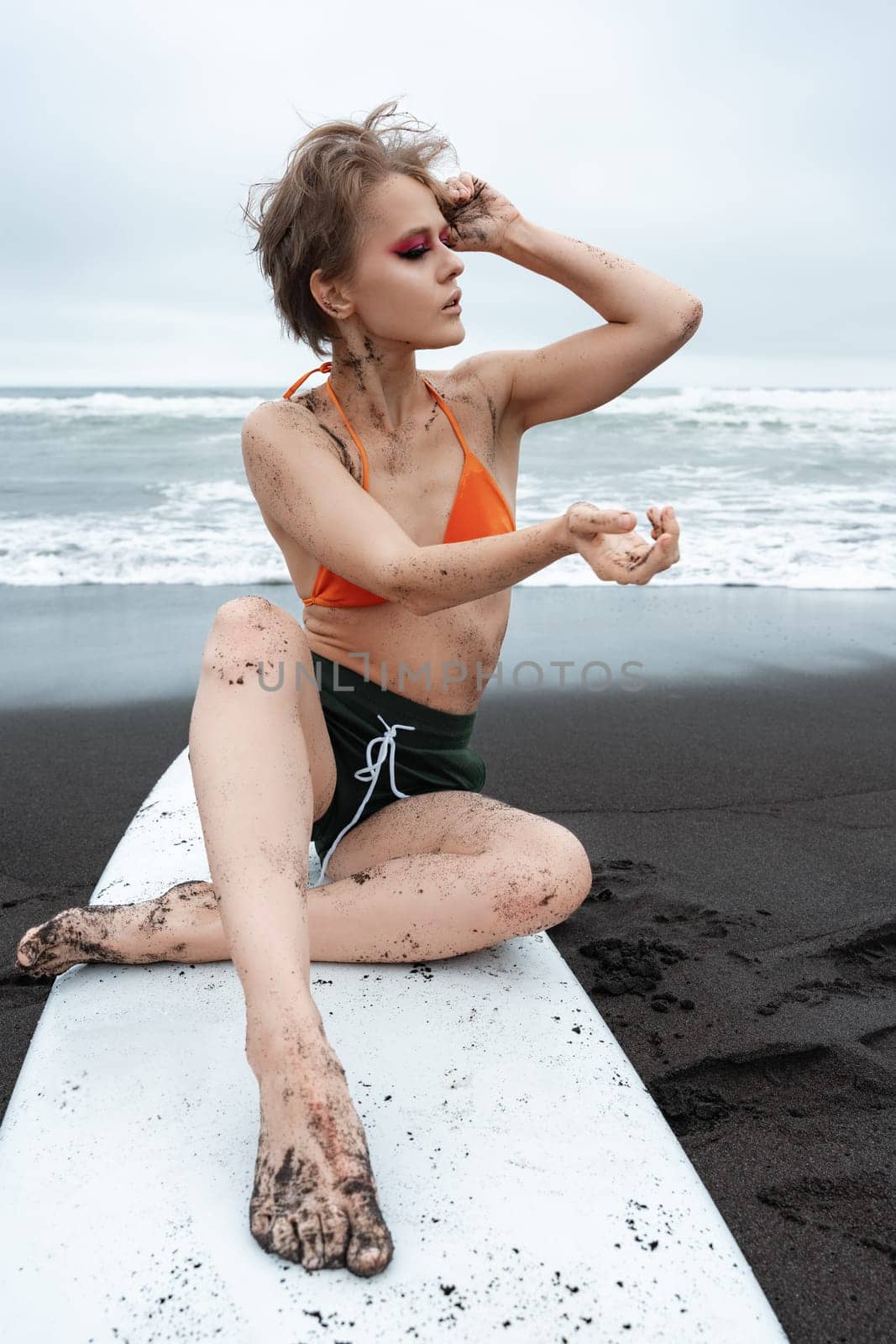 Female surfer sitting on surfboard on black sand beach with sea waves in background. She looks away, exuding authenticity. Woman dressed in bikini top and shorts, and scene is both sexy and athletic