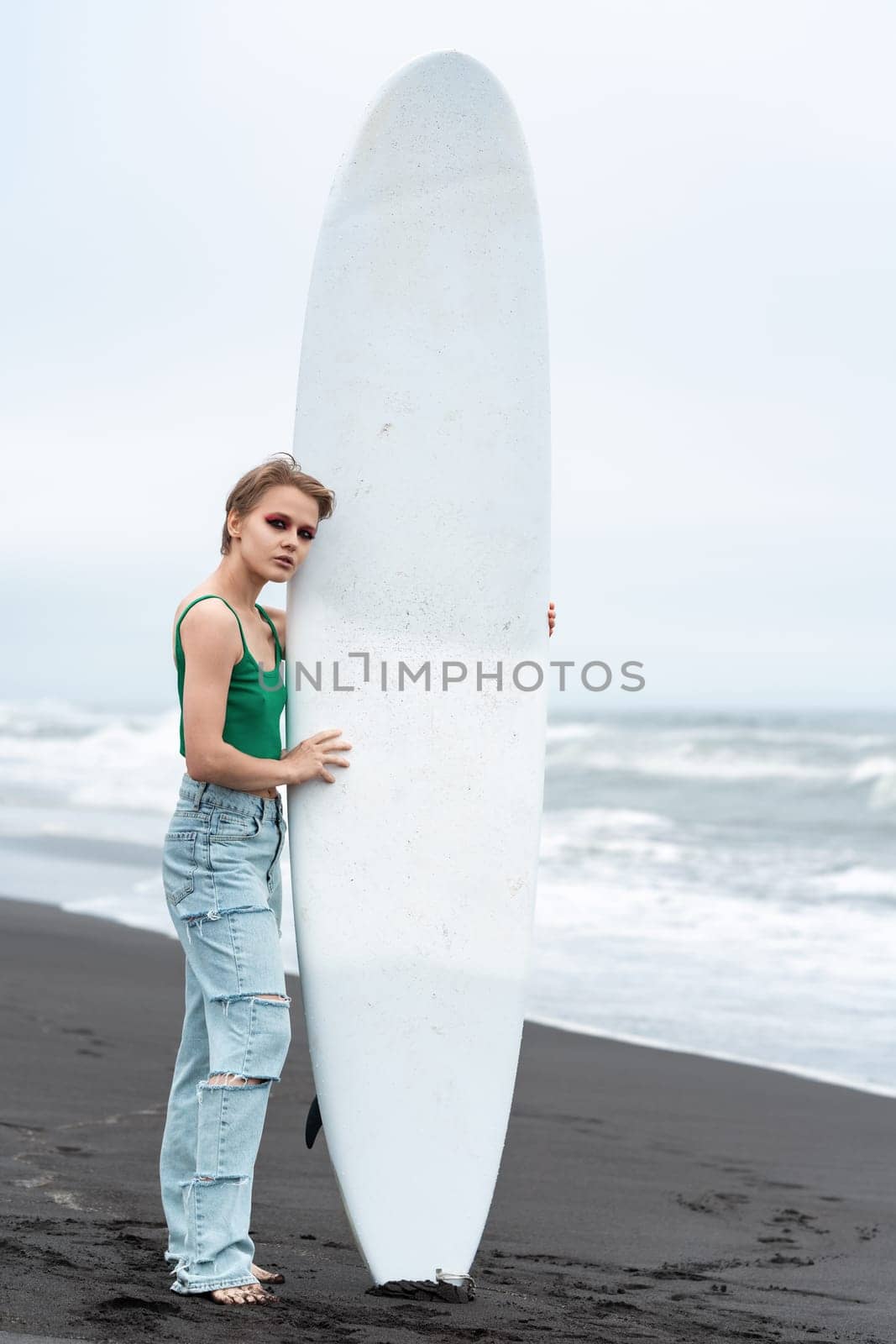 Woman surfer standing on beach, holding surfboard vertically. Fashion model looking at camera, posing against background of breaking ocean waves. Woman wearing casual clothing green top and blue jeans
