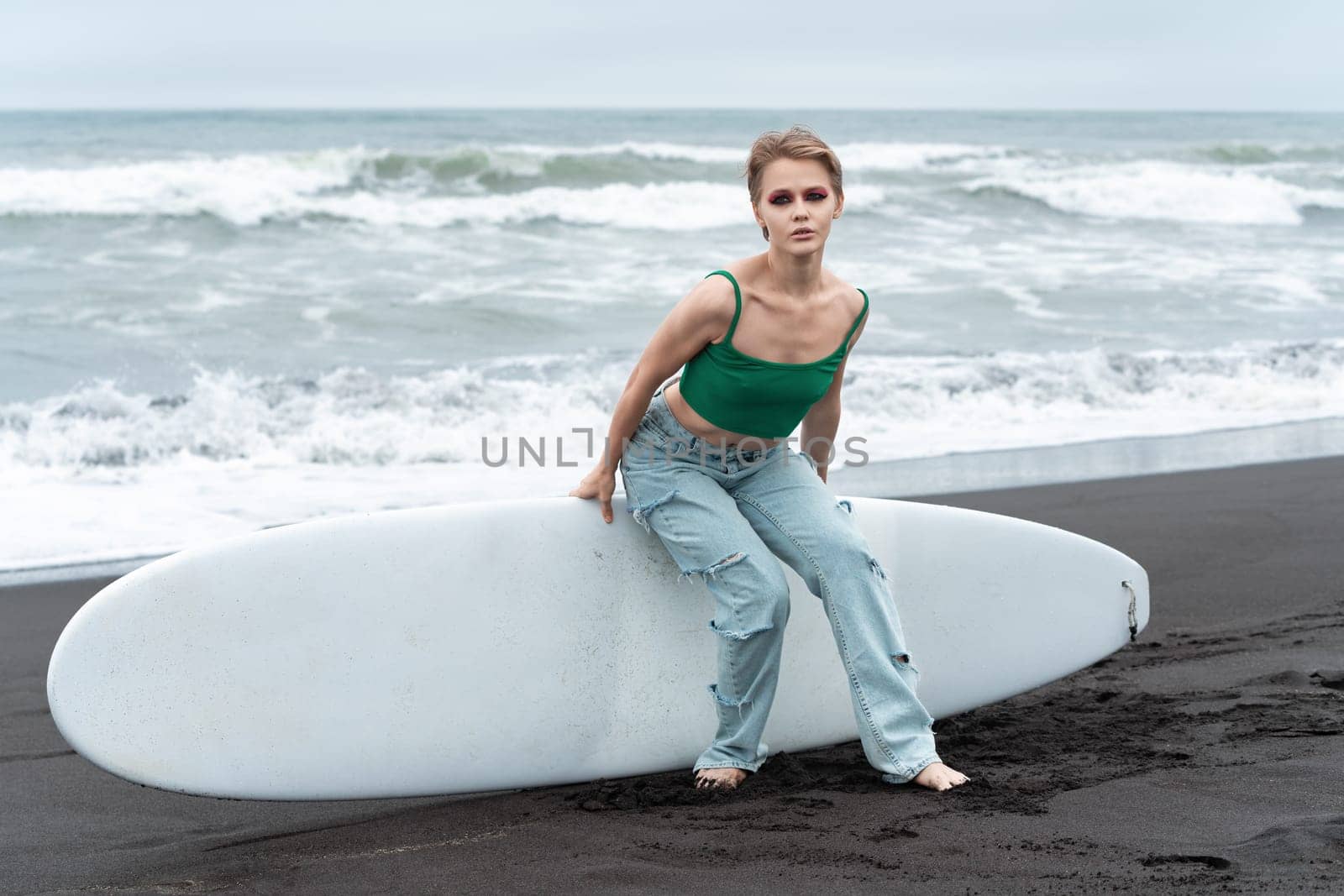 Sensual blonde woman sitting on surf board, lying on sand, against backdrop of pounding waves of Pacific Ocean. Authenticity Caucasian blonde model looking at camera, wearing top and blue jeans