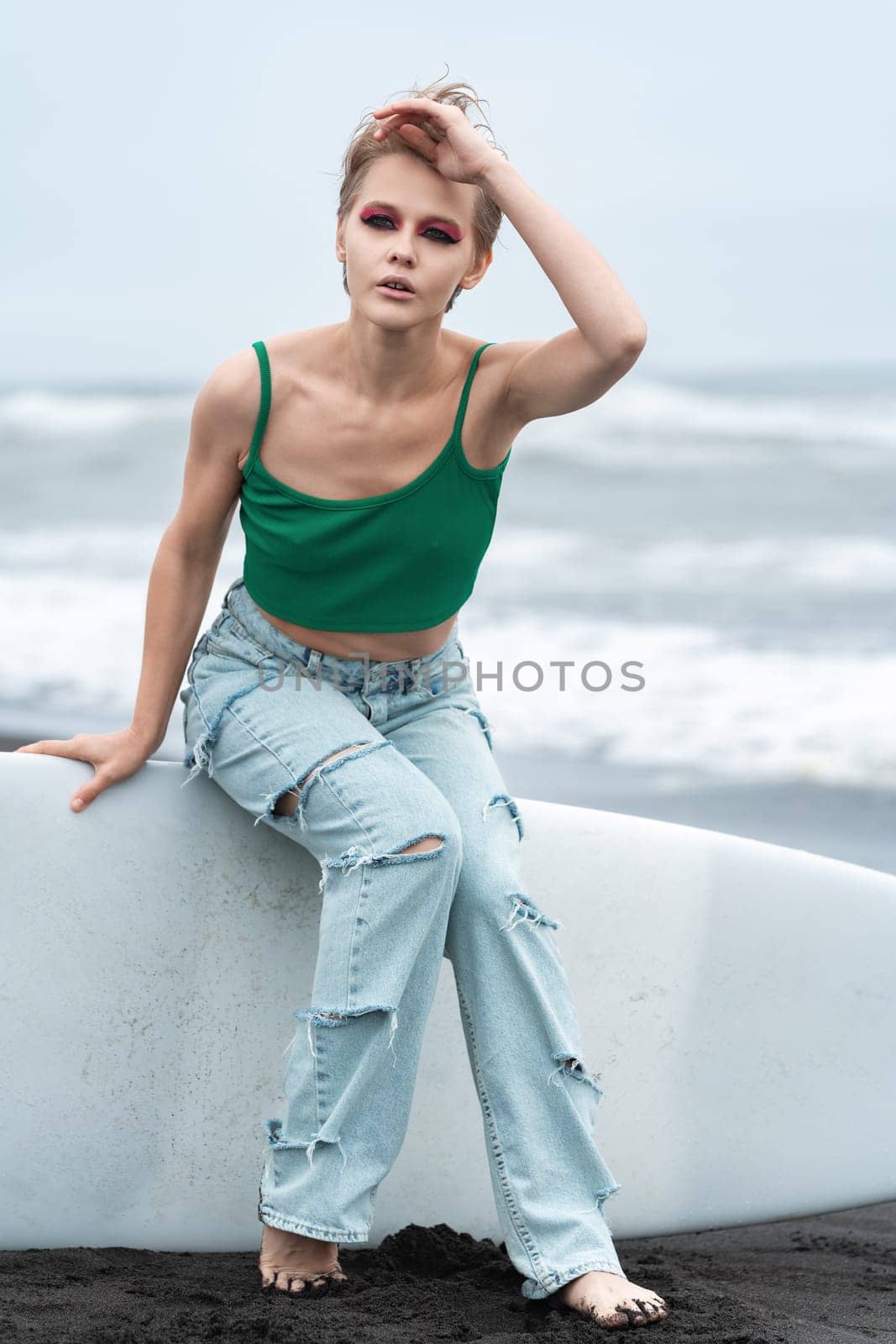 Woman wearing top and blue jeans sitting on surf board lying on sand, on background of sea waves by Alexander-Piragis