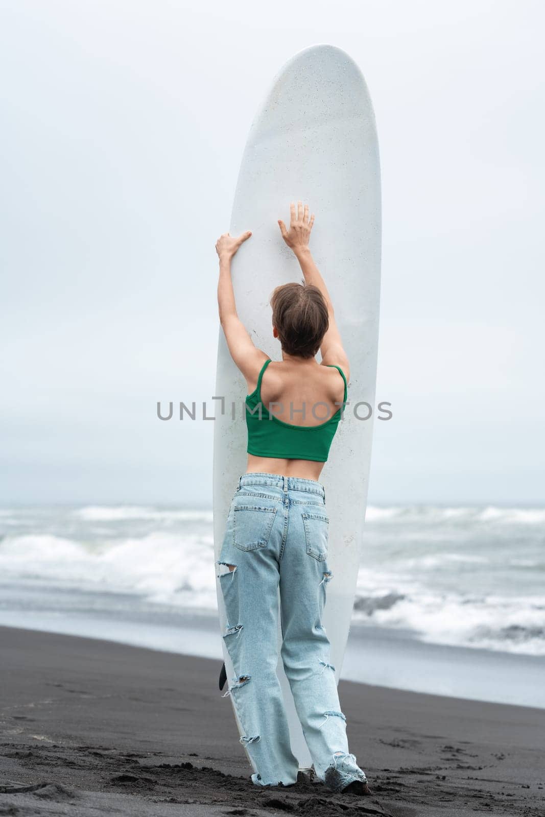 Rear view of woman surfer arms raised holding surfboard vertically, standing on sandy beach, posing by Alexander-Piragis