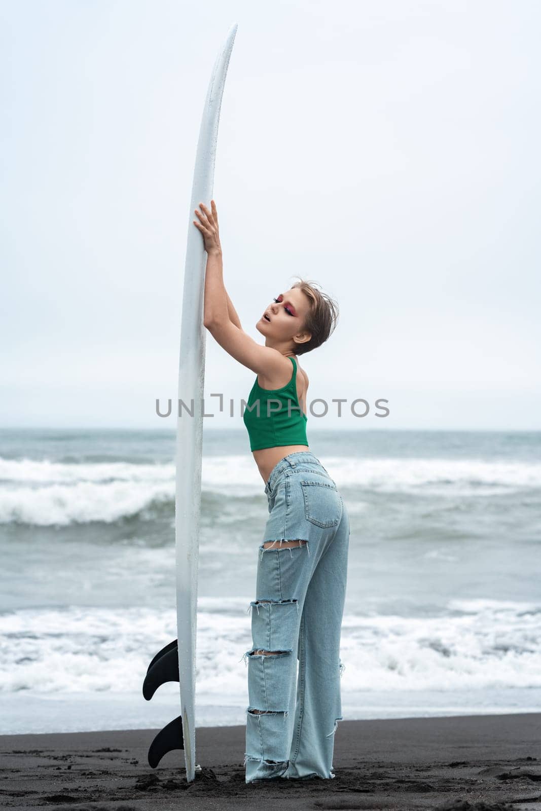 Side view of woman surfer arms raised and holding surfboard vertically. Full length playful female standing on sandy beach, posing on background of ocean waves. Hipster woman wearing casual clothing