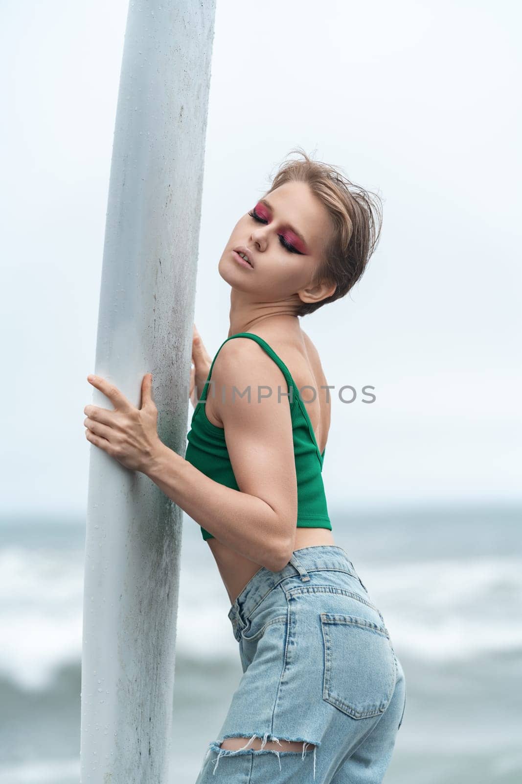 Portrait of sensual beautiful woman with closed eyes holding white surfboard with both hands, posing sexy. Fashion model with short hair and bright makeup wearing casual clothing - top and blue jeans